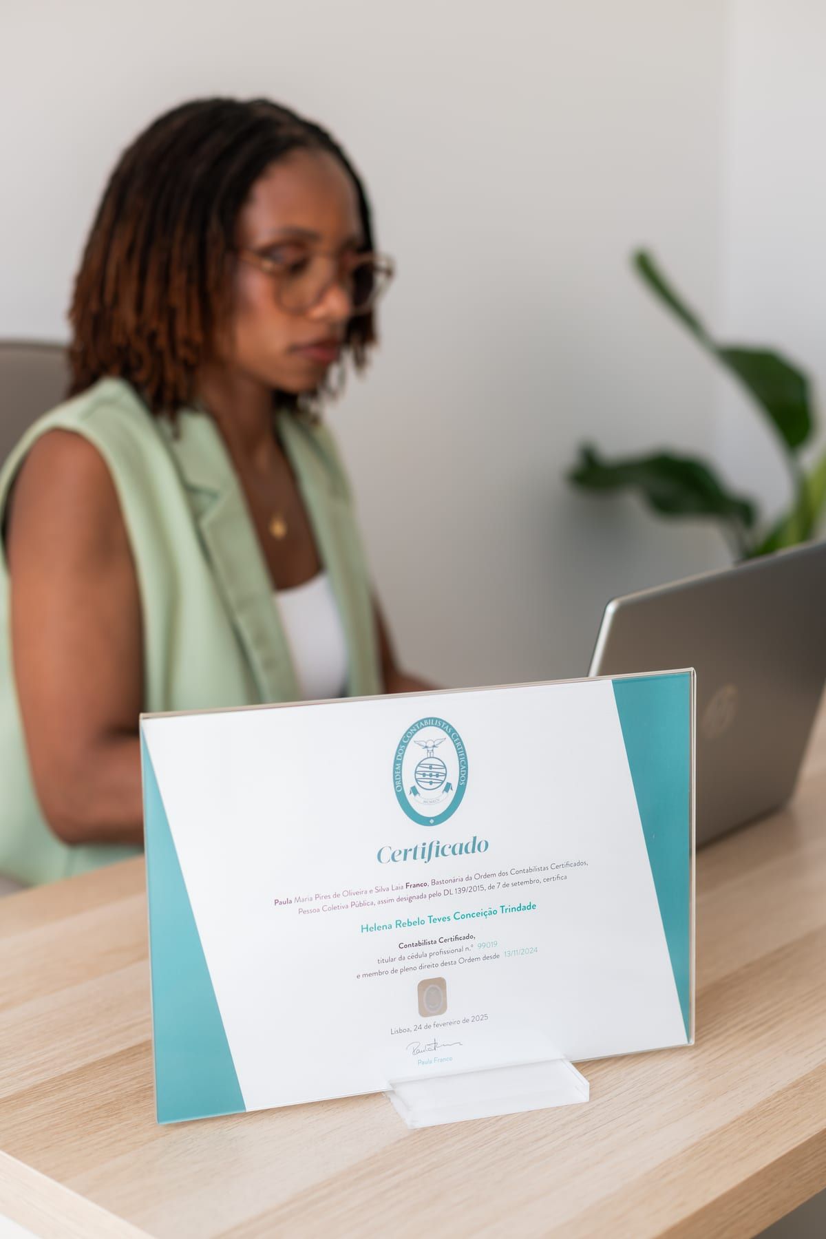 Certificado profissional de Helena Trindade sobre a secretária, com a Helena a trabalhar ao fundo